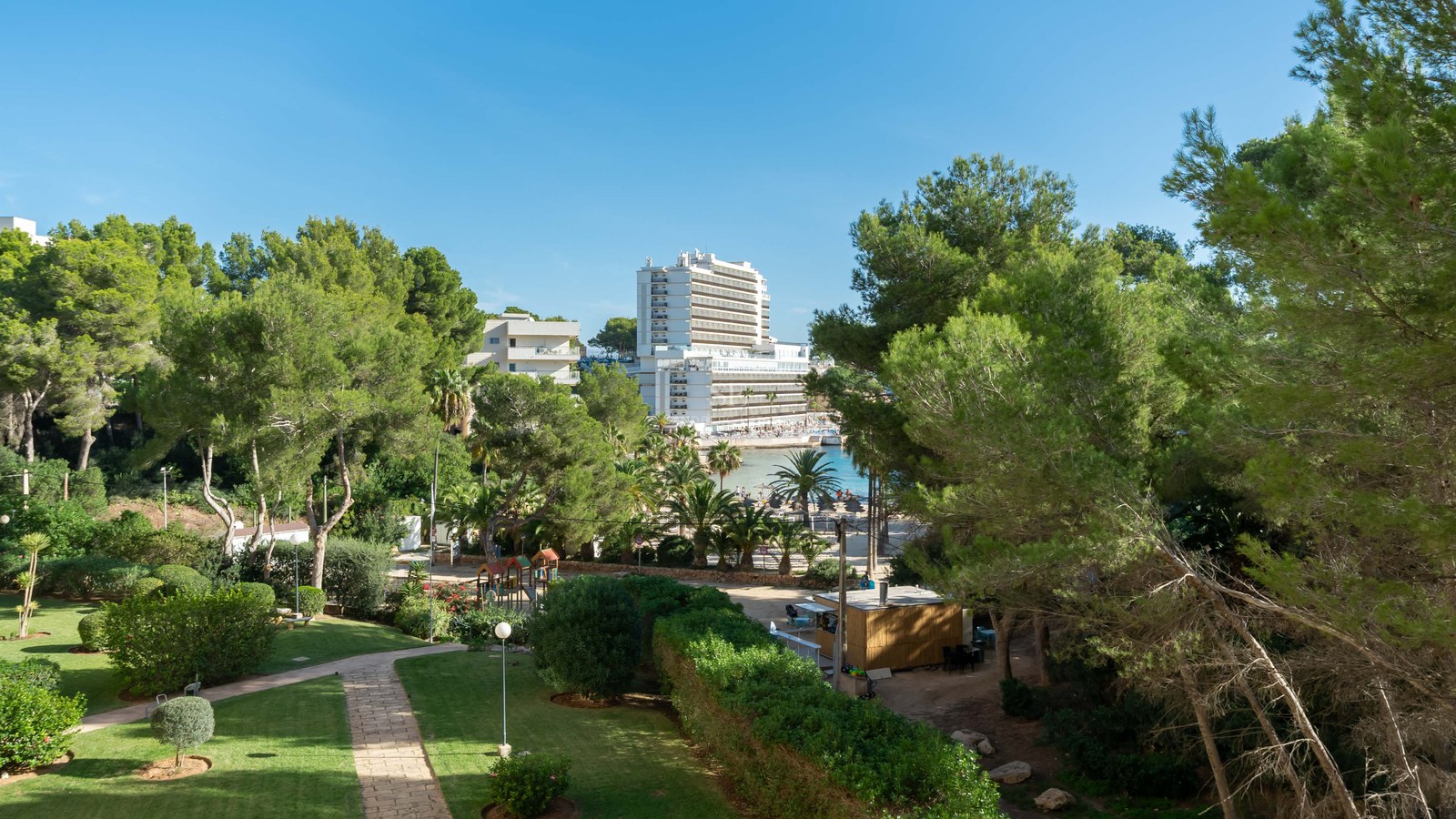 Jardín verde cuidado con caminos de piedra, pinos y un edificio alto con vistas a una bahía bajo un cielo azul despejado.