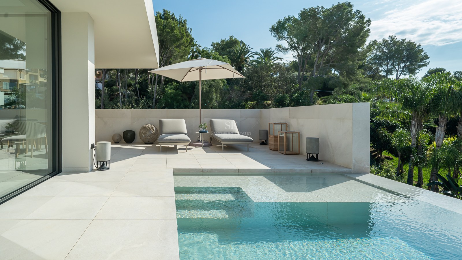 Piscine à débordement moderne avec terrasse en pierre blanche, chaises longues, parasol et arrière-plan de verdure.