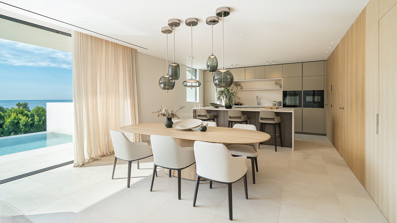 Salle à manger contemporaine avec table en bois et suspensions en verre, surplombant une piscine et la mer.