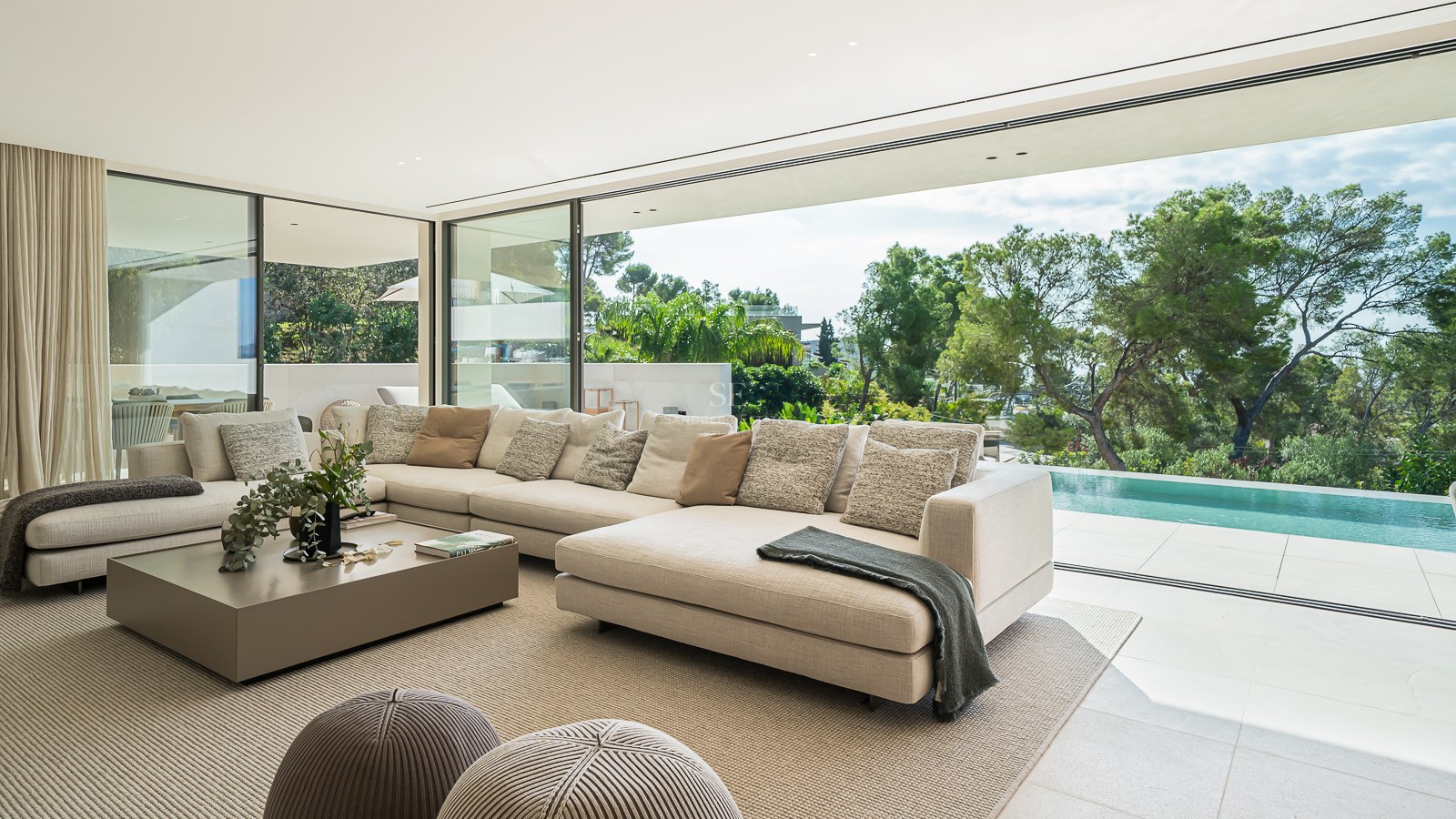 Un salon moderne et lumineux avec un grand canapé d'angle crème donnant sur une piscine privée et des arbres.