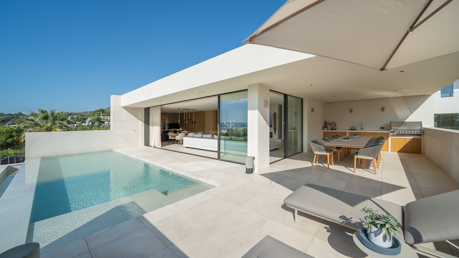 Terrasse de villa moderne avec piscine à débordement, chaises longues, salle à manger extérieure et espace de vie ouvert.