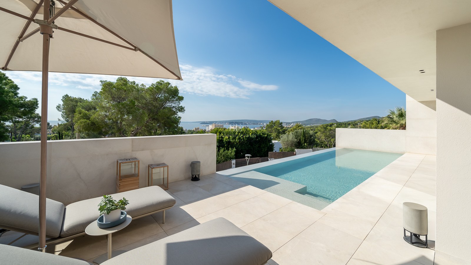 Chaises longues sur une terrasse moderne en pierre beige à côté d'une piscine offrant une vue sur la mer et la végétation.