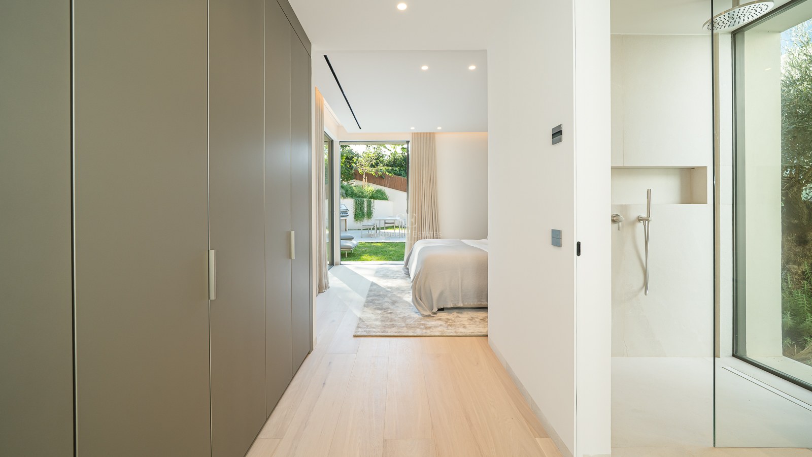 Couloir avec placards menant à une chambre moderne avec douche vitrée et accès direct à un jardin ensoleillé.