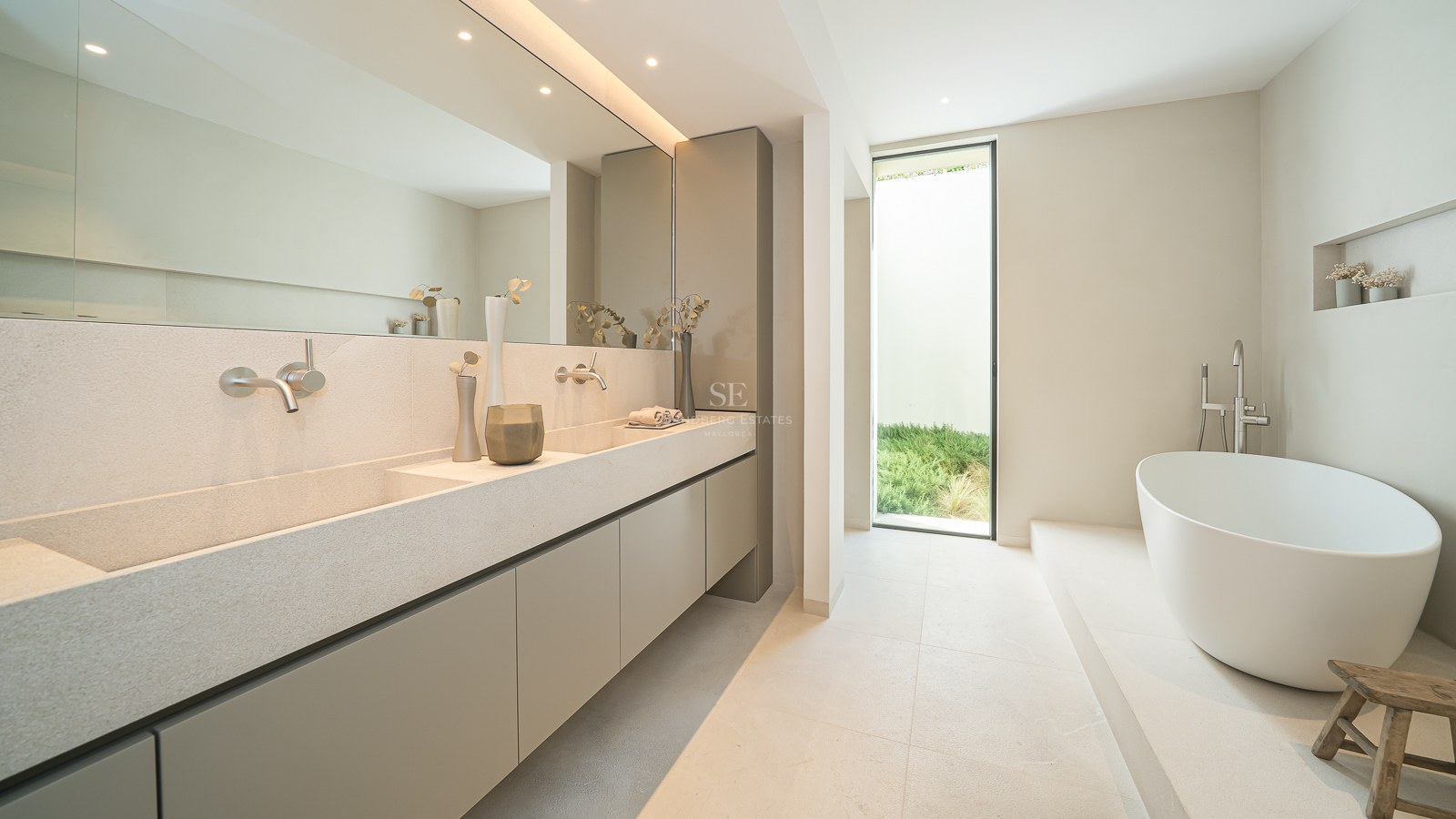 Salle de bain minimaliste avec double vasque en pierre, grand miroir, baignoire blanche et fenêtre sur le jardin.