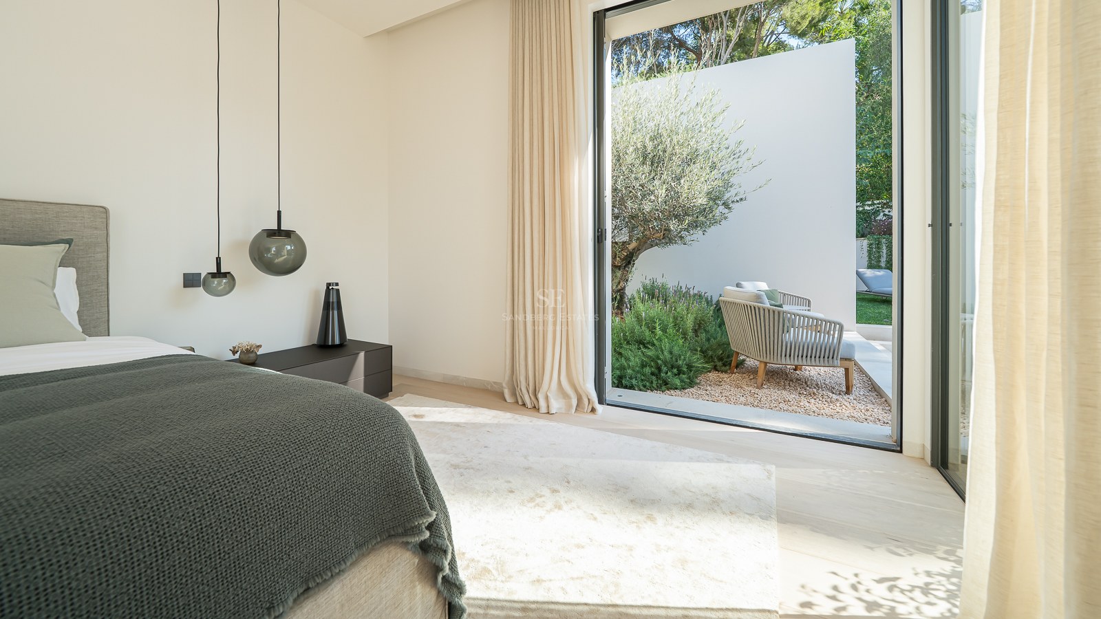 Chambre moderne avec un grand lit, des suspensions design et des baies vitrées ouvrant sur un jardin avec un olivier.