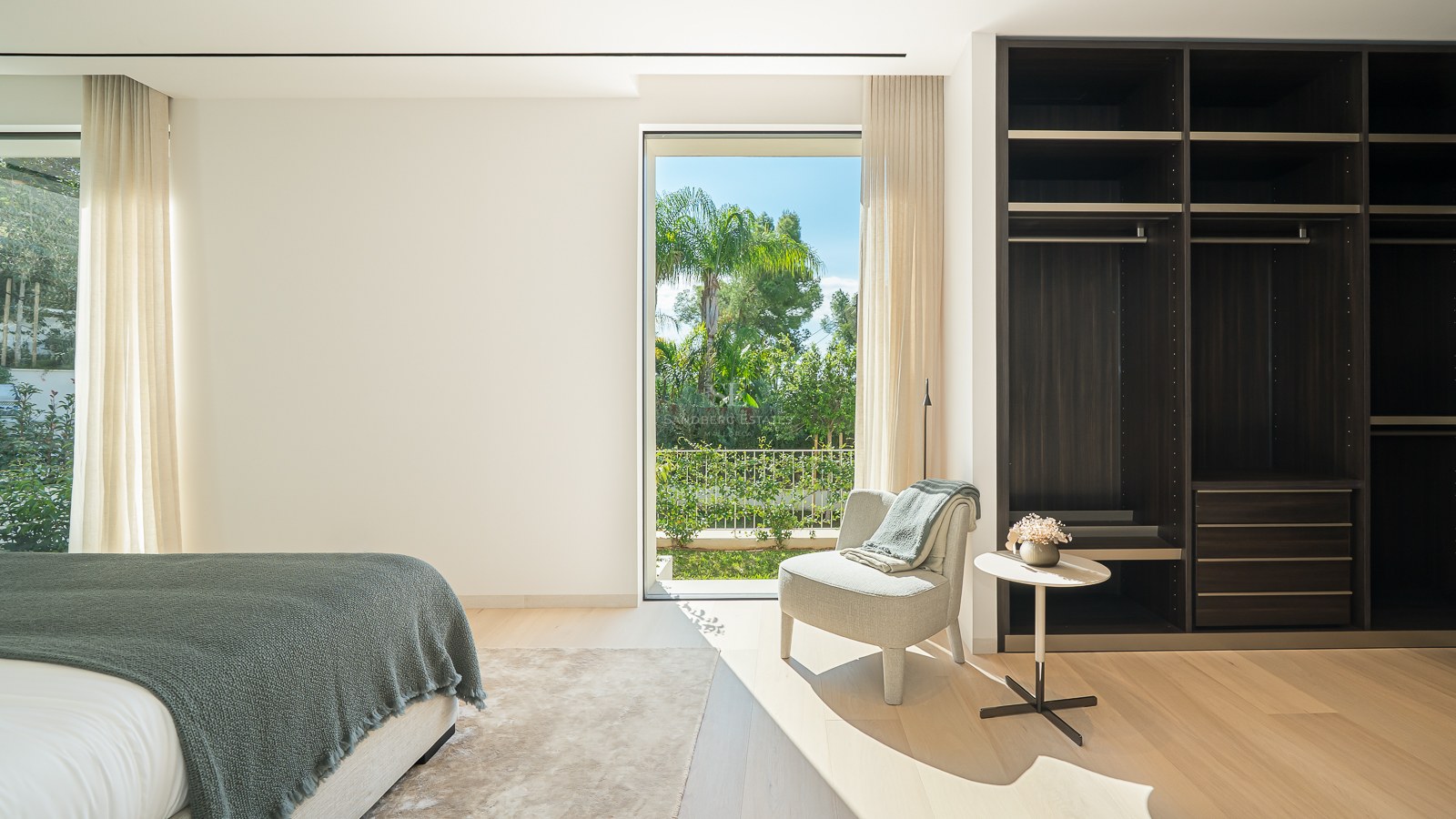 Chambre lumineuse avec un grand dressing en bois sombre, un fauteuil et une vue sur un jardin tropical.