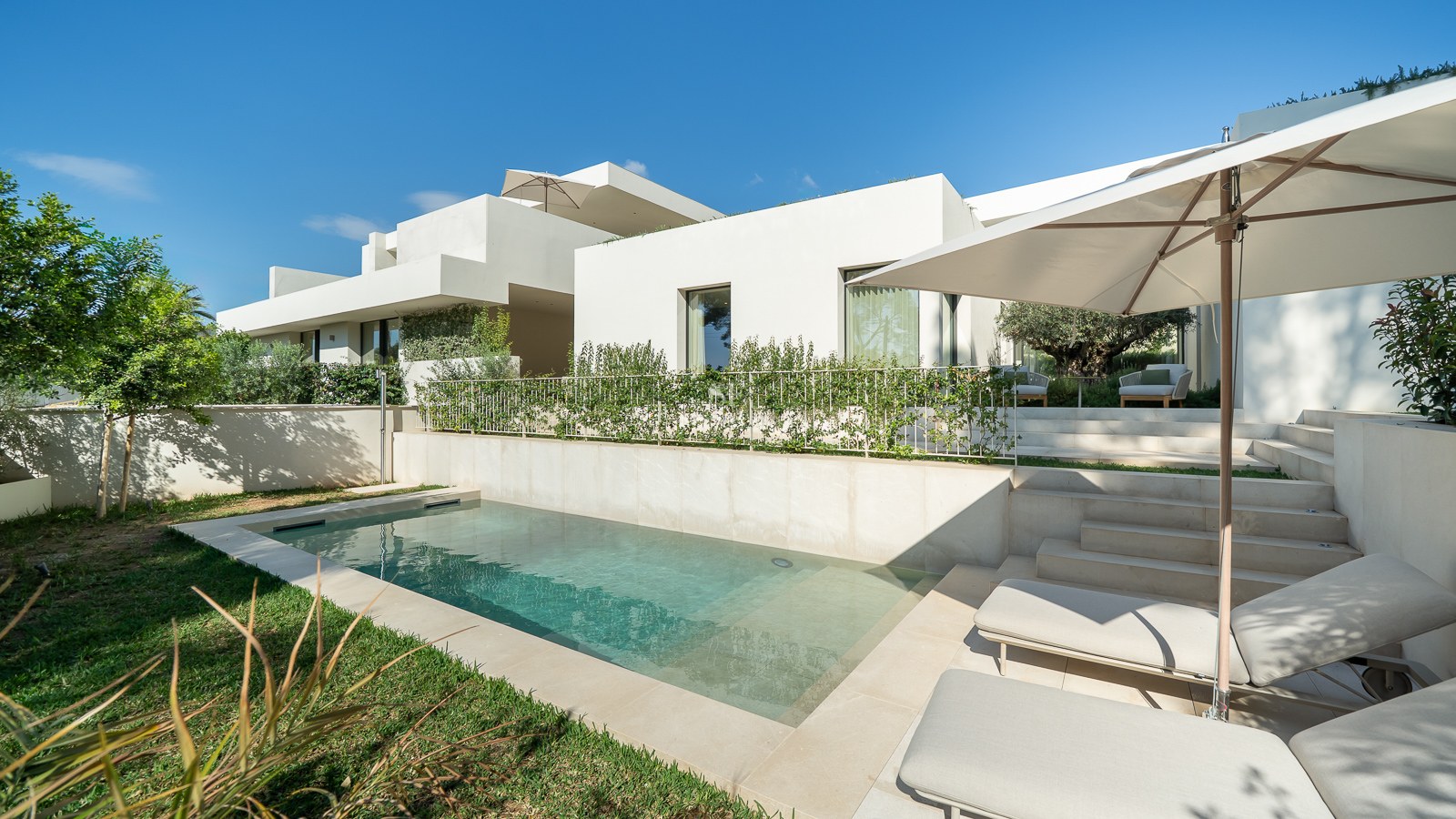 Villa blanche moderne avec piscine rectangulaire, patio en pierre, bains de soleil et jardin verdoyant sous un ciel bleu.