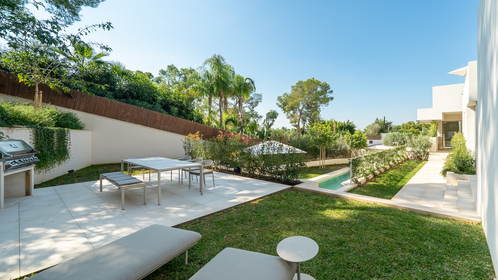 Terrasse moderne avec coin repas, barbecue et piscine entourée d'une végétation luxuriante et de palmiers.
