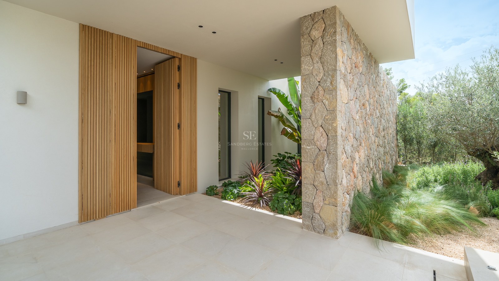 Entrée d'une maison moderne avec porte en bois à lattes, mur en pierre naturelle et plantes tropicales.