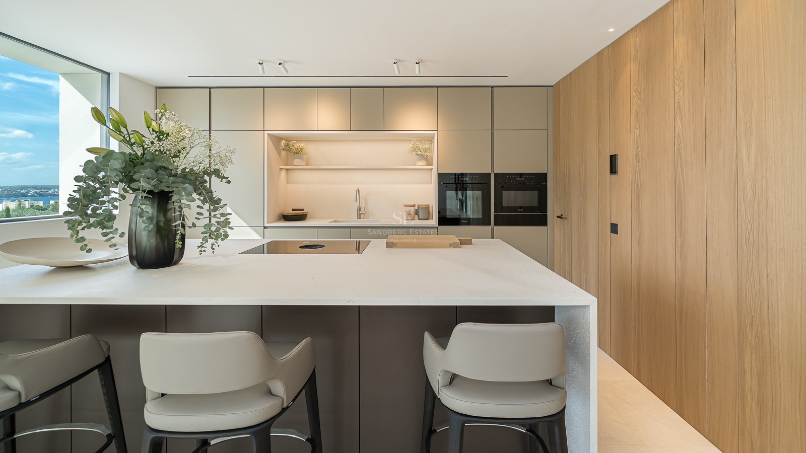 Îlot de cuisine moderne avec tabourets, appareils intégrés, mur en bois et fenêtre panoramique sur l'océan.