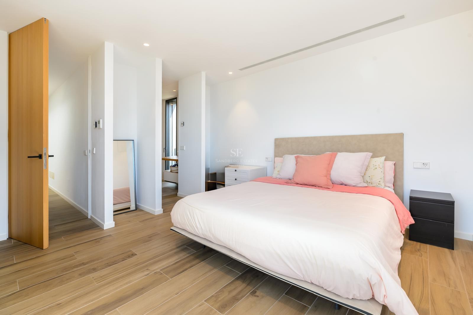 Dormitorio luminoso y moderno con una cama grande, cojines rosas, suelos de madera clara y paredes blancas.