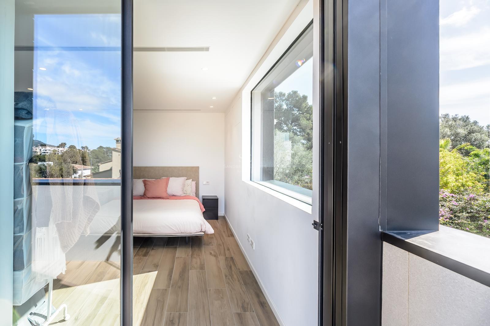 Dormitorio moderno con suelos de madera, una cama con cojines rosas y grandes ventanales con vistas a árboles verdes.