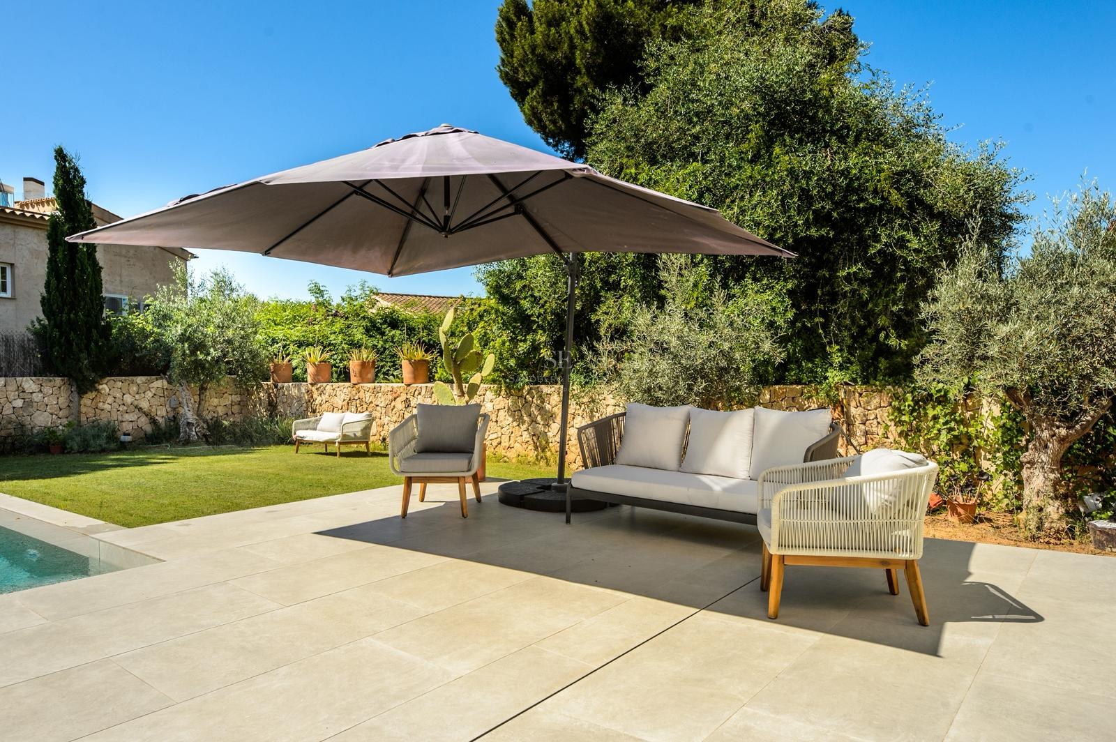 Terraza soleada con sofá gris moderno, sillones y sombrilla grande junto a una piscina y jardín.
