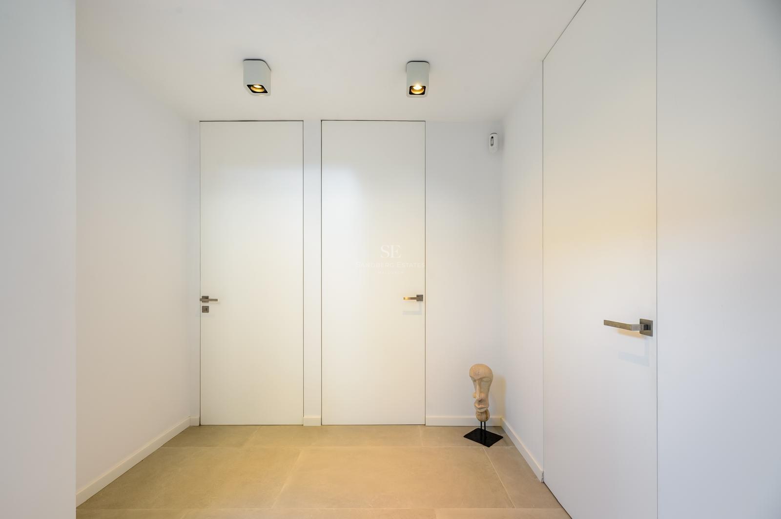 Pasillo minimalista con puertas blancas integradas, suelo de piedra clara y una pequeña escultura decorativa.