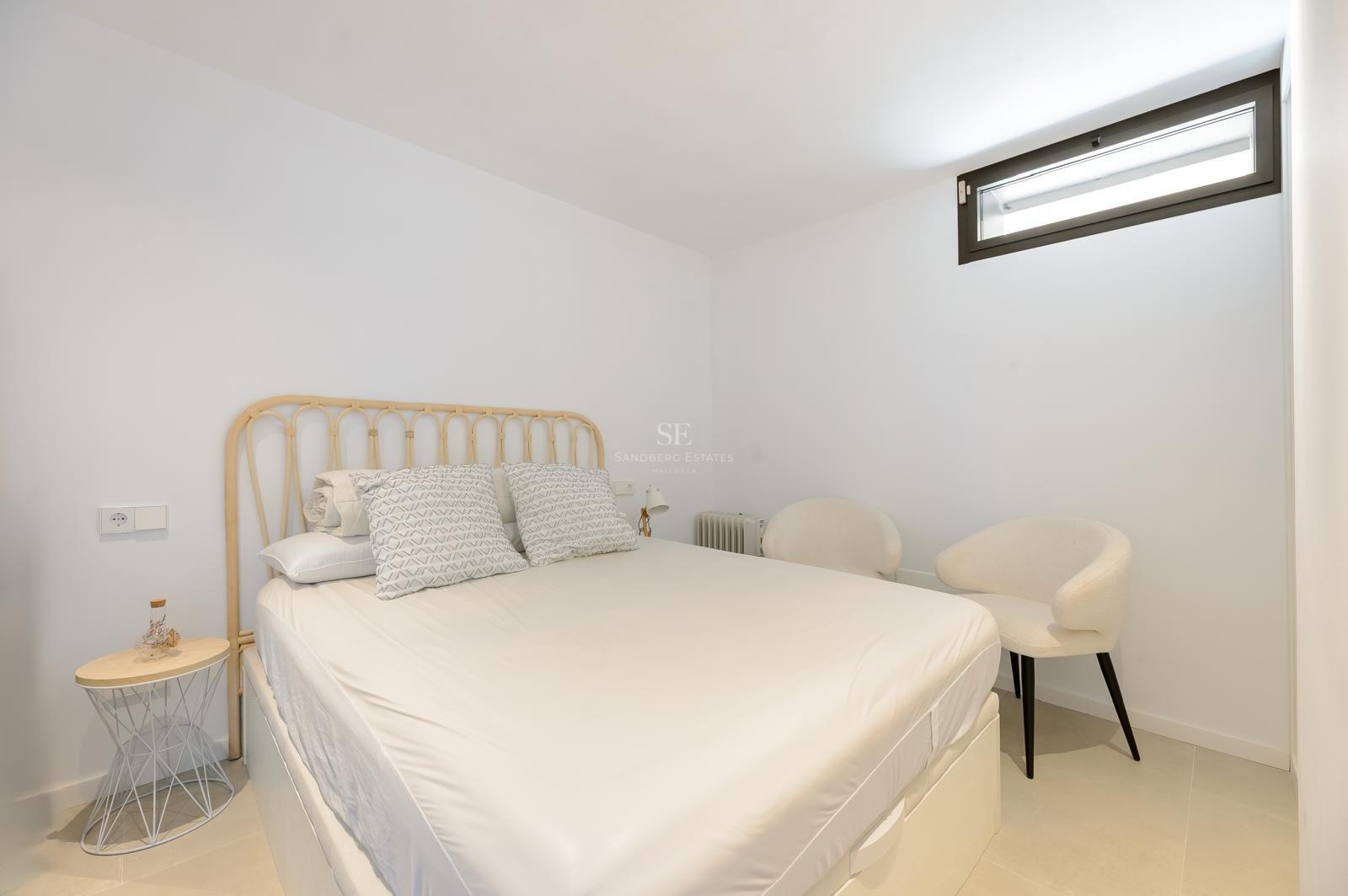 Dormitorio luminoso con cabecero de madera clara, cama blanca y dos sillas modernas junto a una ventana pequeña.