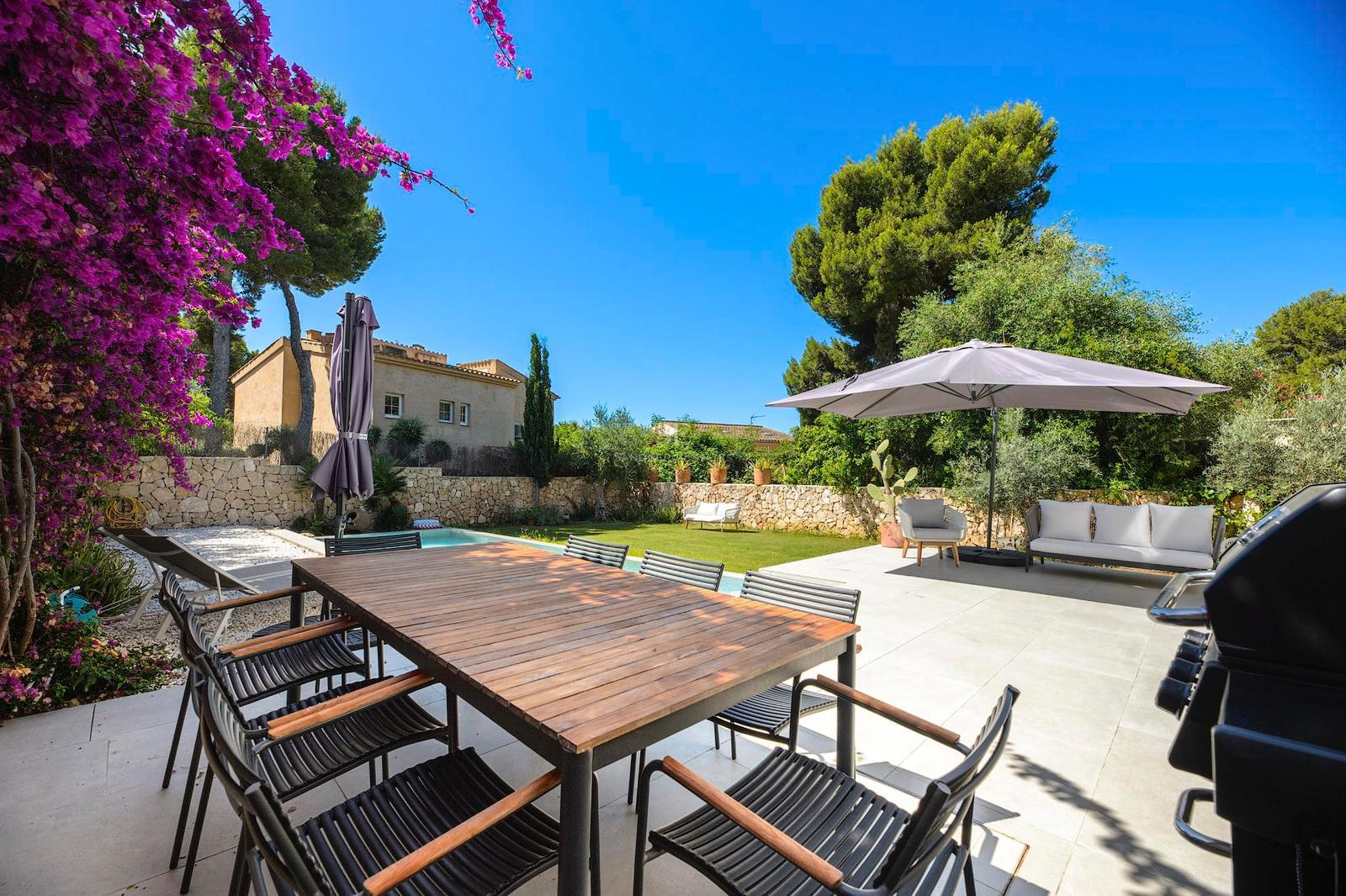Mesa de comedor de madera en terraza soleada con vistas a un jardín impecable y piscina privada.