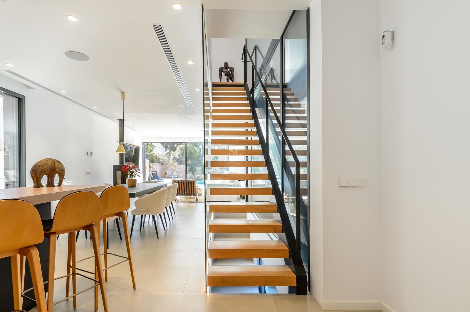 Interior moderno con escalera flotante de madera, barandilla de cristal y salón luminoso al fondo.