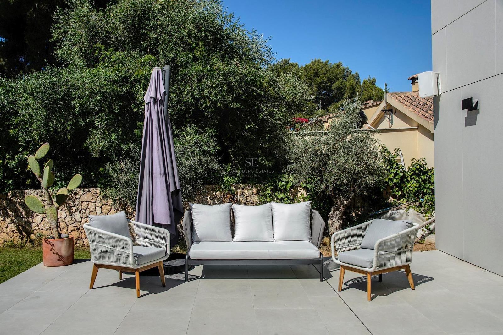 Sofá y sillones de cuerda trenzada gris en terraza de piedra rodeada de vegetación mediterránea bajo cielo azul.