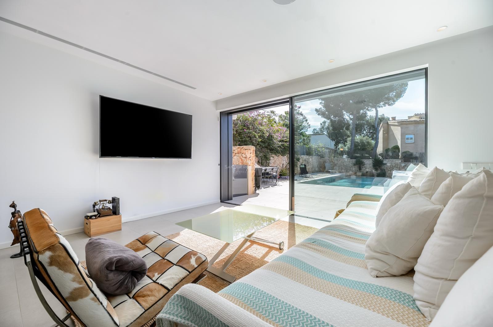 Salón luminoso con sofá a rayas, televisor grande y puertas de cristal que dan a una terraza con piscina.