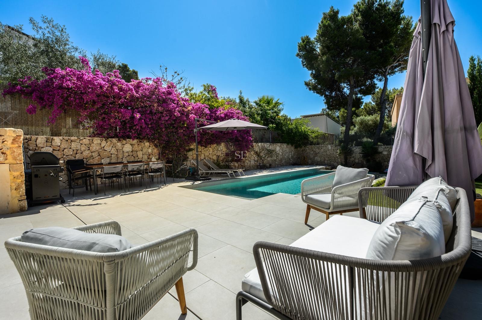Zona de piscina privada con asientos de exterior, mesa de comedor, barbacoa y buganvillas púrpuras en muro de piedra.