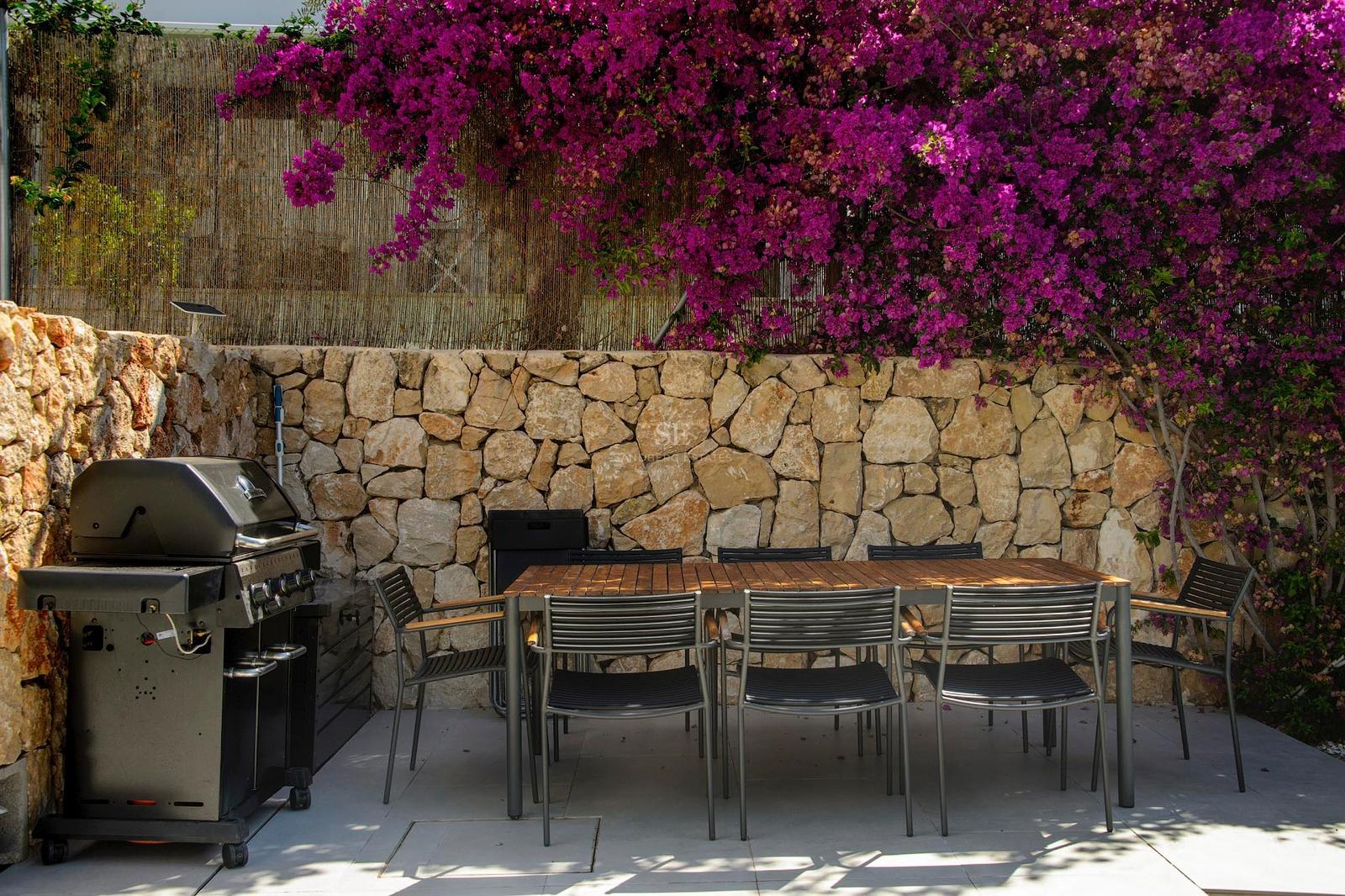 Terraza con mesa de madera, sillas negras, barbacoa y muro de piedra con flores de buganvilla rosa.