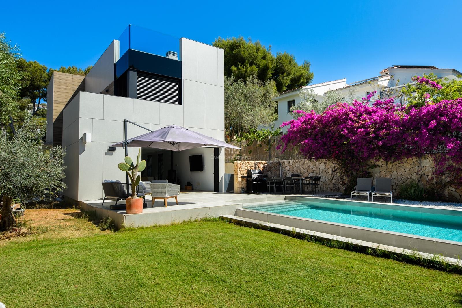 Exterior de villa moderna con piscina azul, césped verde y buganvilla morada contra un muro de piedra.
