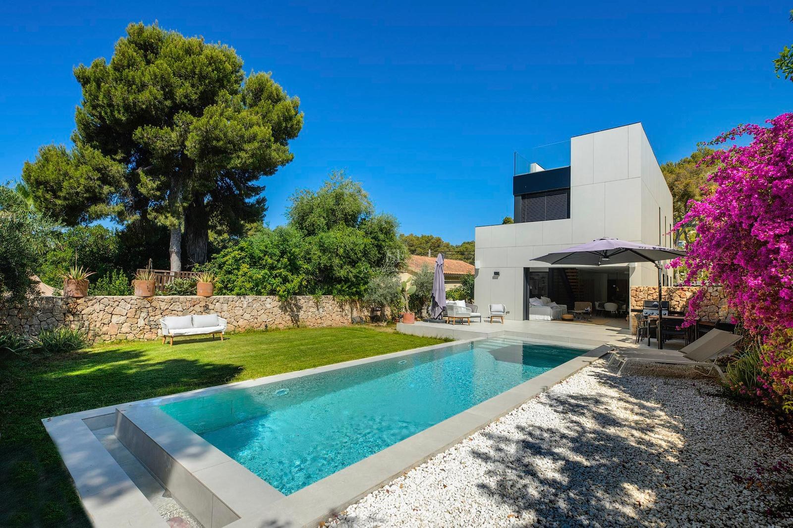Piscina rectangular rodeada de césped, grava blanca y una villa blanca moderna bajo un cielo azul despejado.