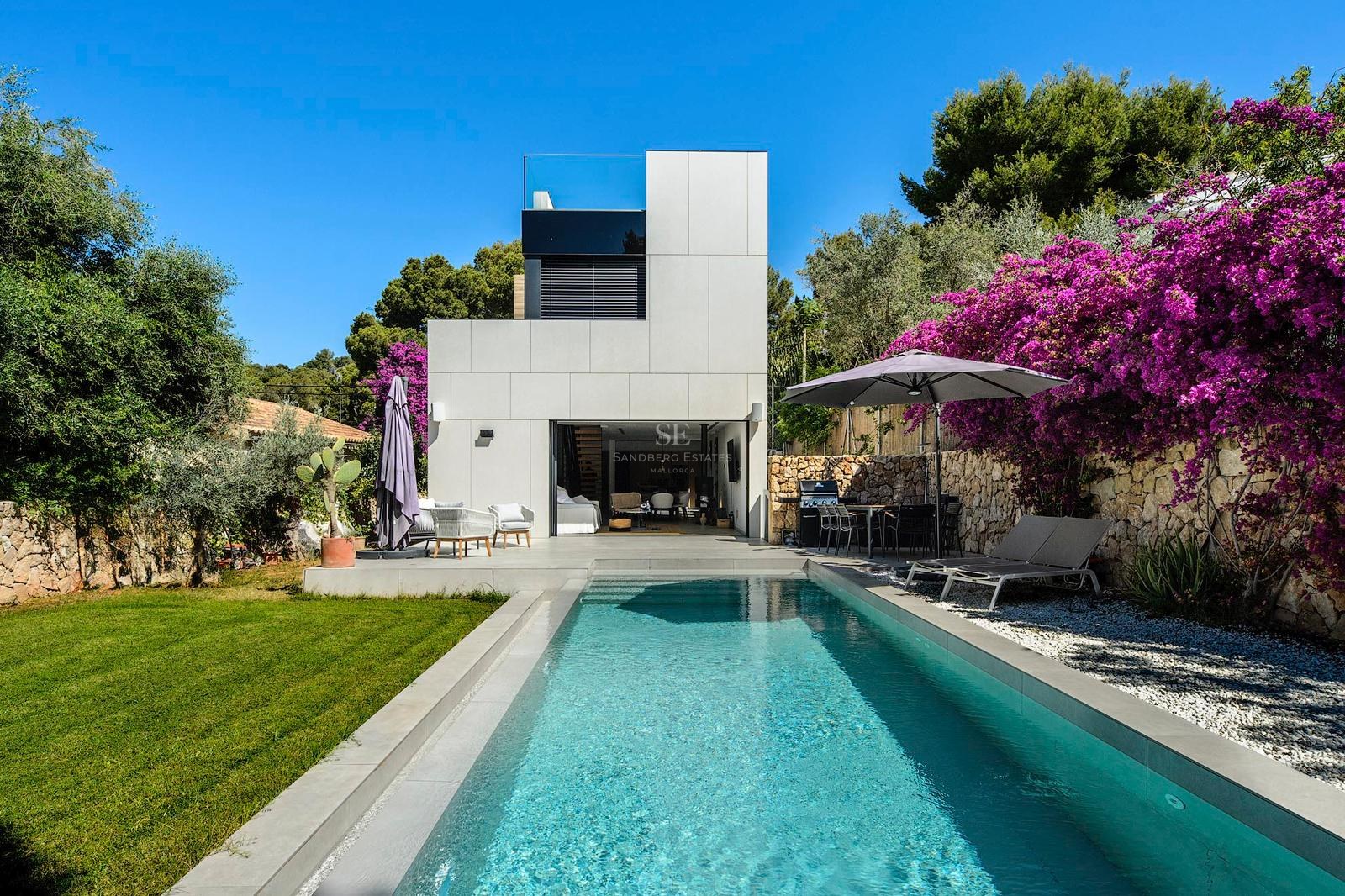 Villa blanca moderna con una piscina rectangular, césped verde y buganvillas púrpuras bajo un cielo azul despejado.