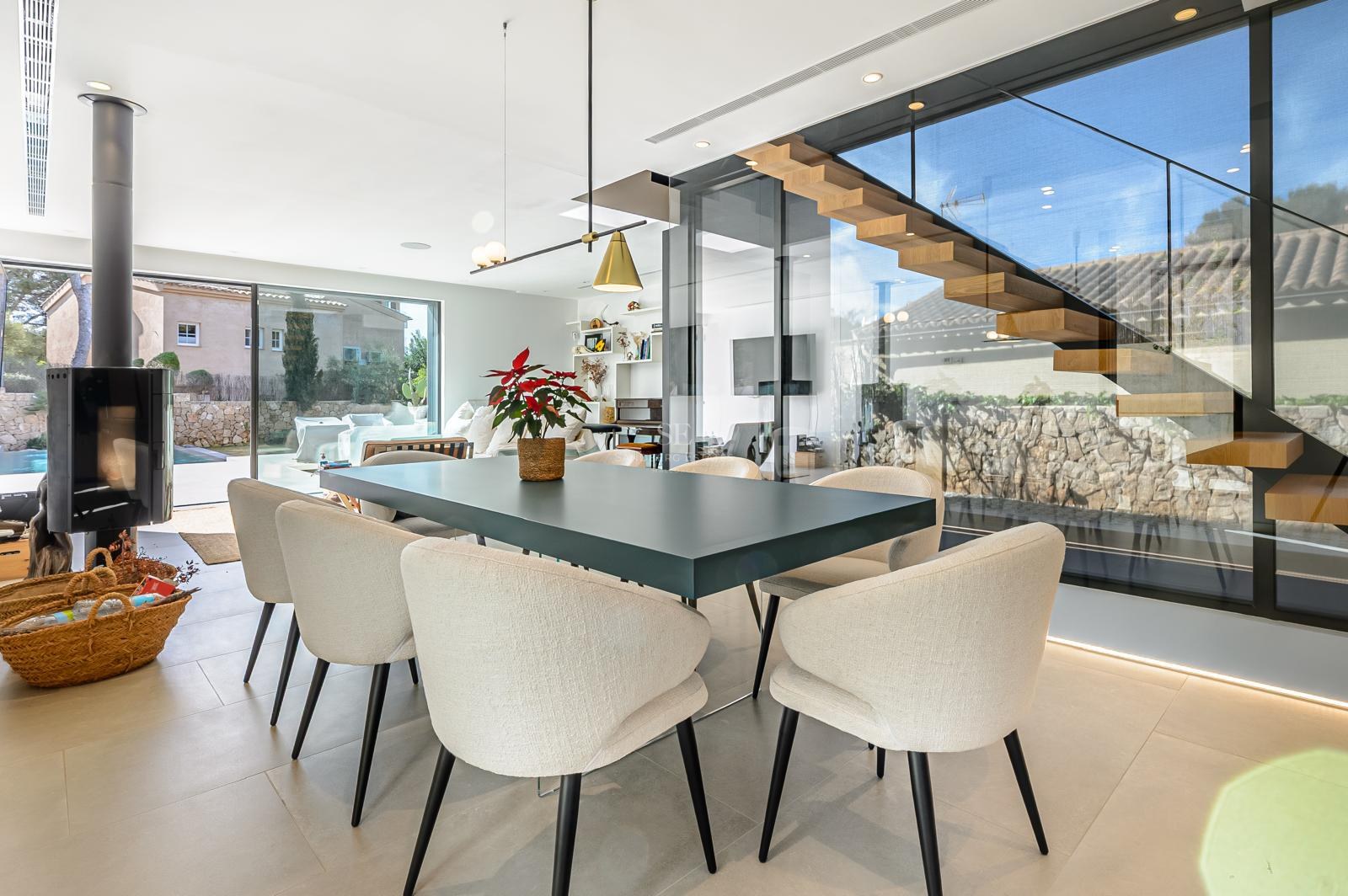 Comedor contemporáneo con mesa oscura, sillas beige, escalera de madera flotante y grandes ventanales de cristal.