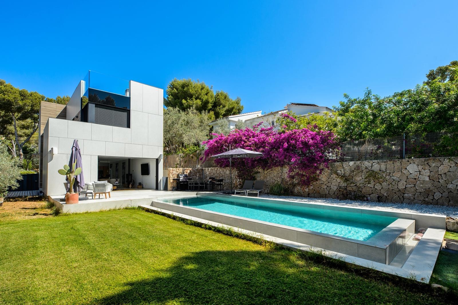 Piscina rectangular moderna junto a una villa blanca con buganvillas púrpuras y césped verde bajo un cielo azul.