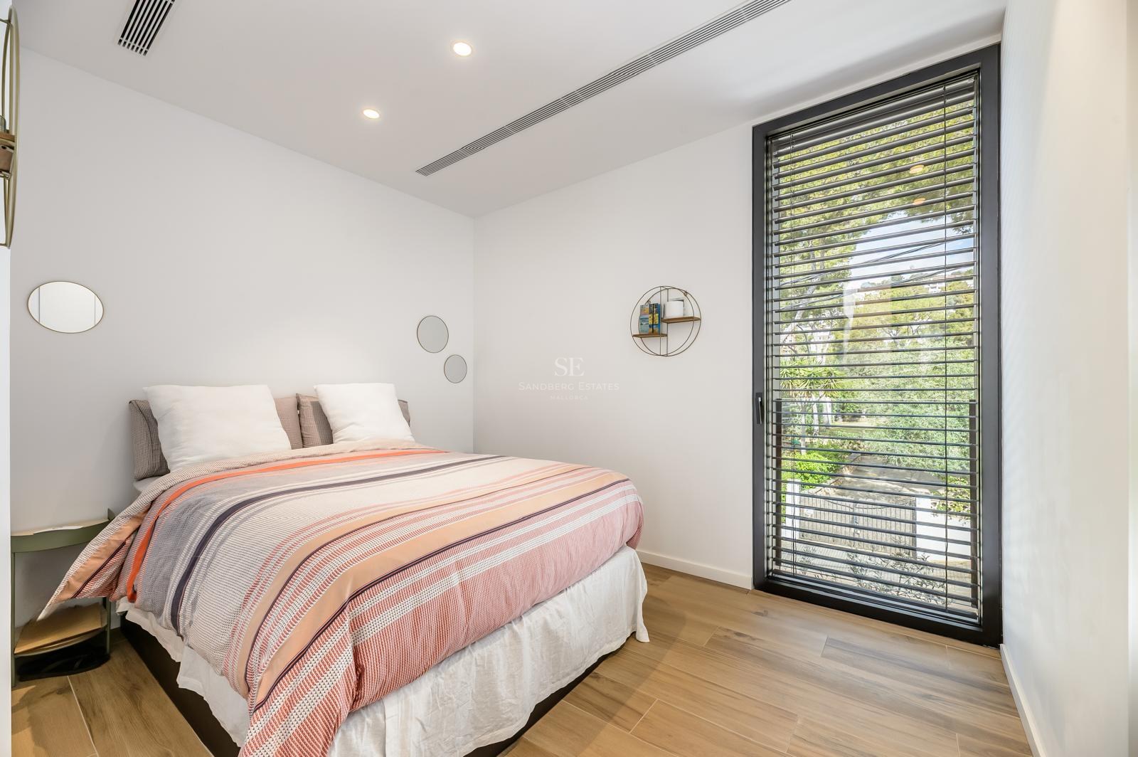 Dormitorio luminoso con cama a rayas, suelos de madera y un gran ventanal con persianas que da a árboles verdes.