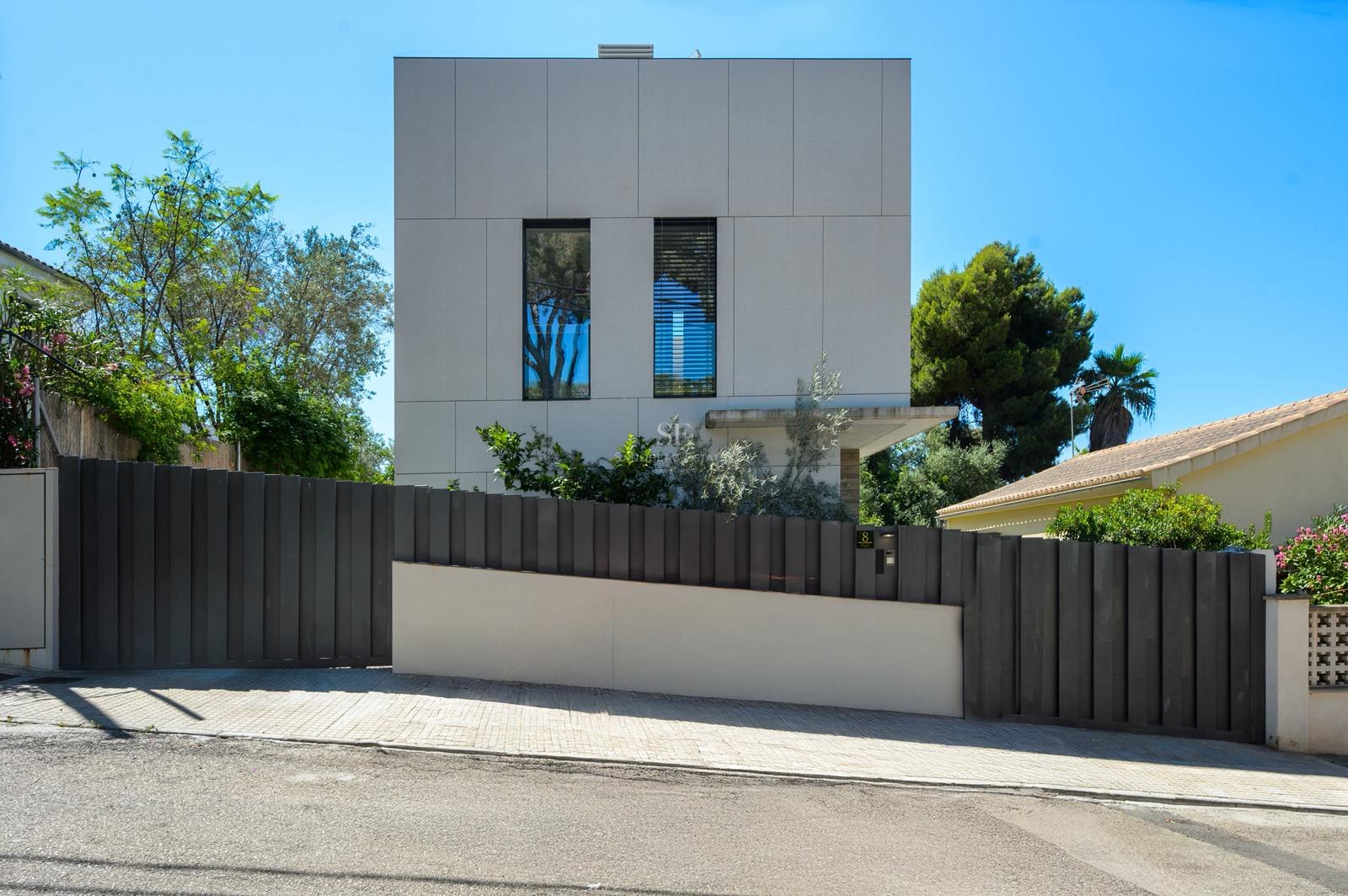 Vista exterior de una casa cúbica blanca moderna con una valla metálica gris oscuro y árboles verdes bajo un cielo azul.