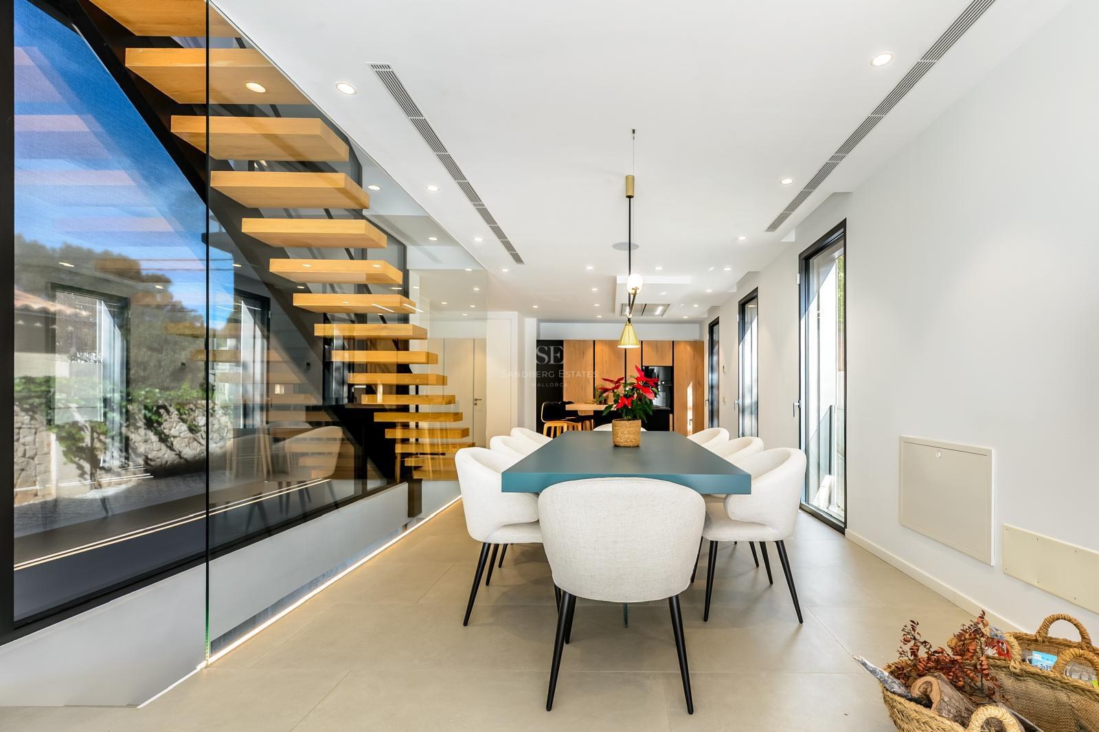 Comedor moderno con mesa azul, sillas blancas y una escalera de madera flotante tras un panel de cristal.