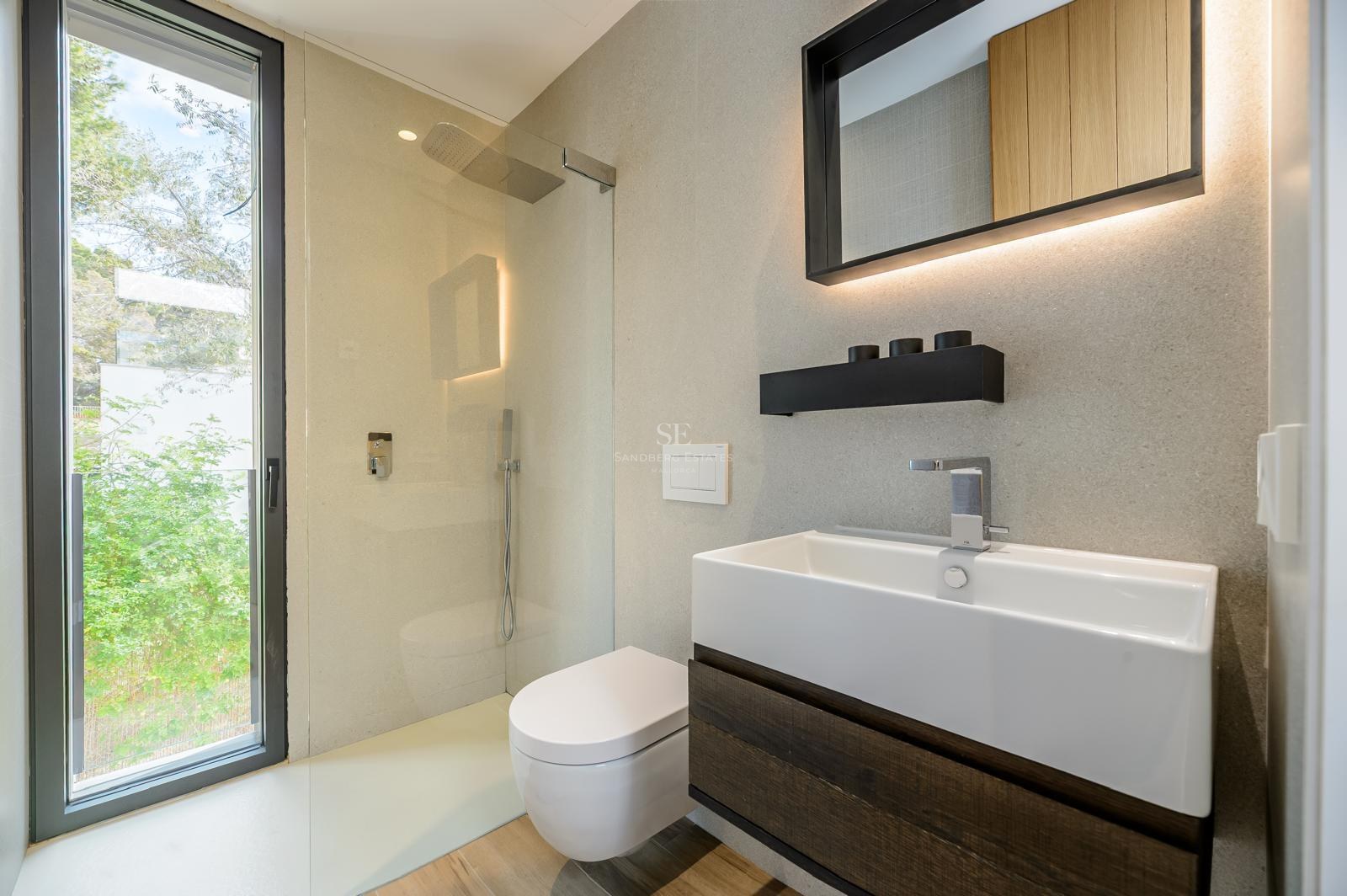 Baño moderno con lavabo blanco, mueble de madera oscura, ducha de cristal y ventanal con vistas al jardín.