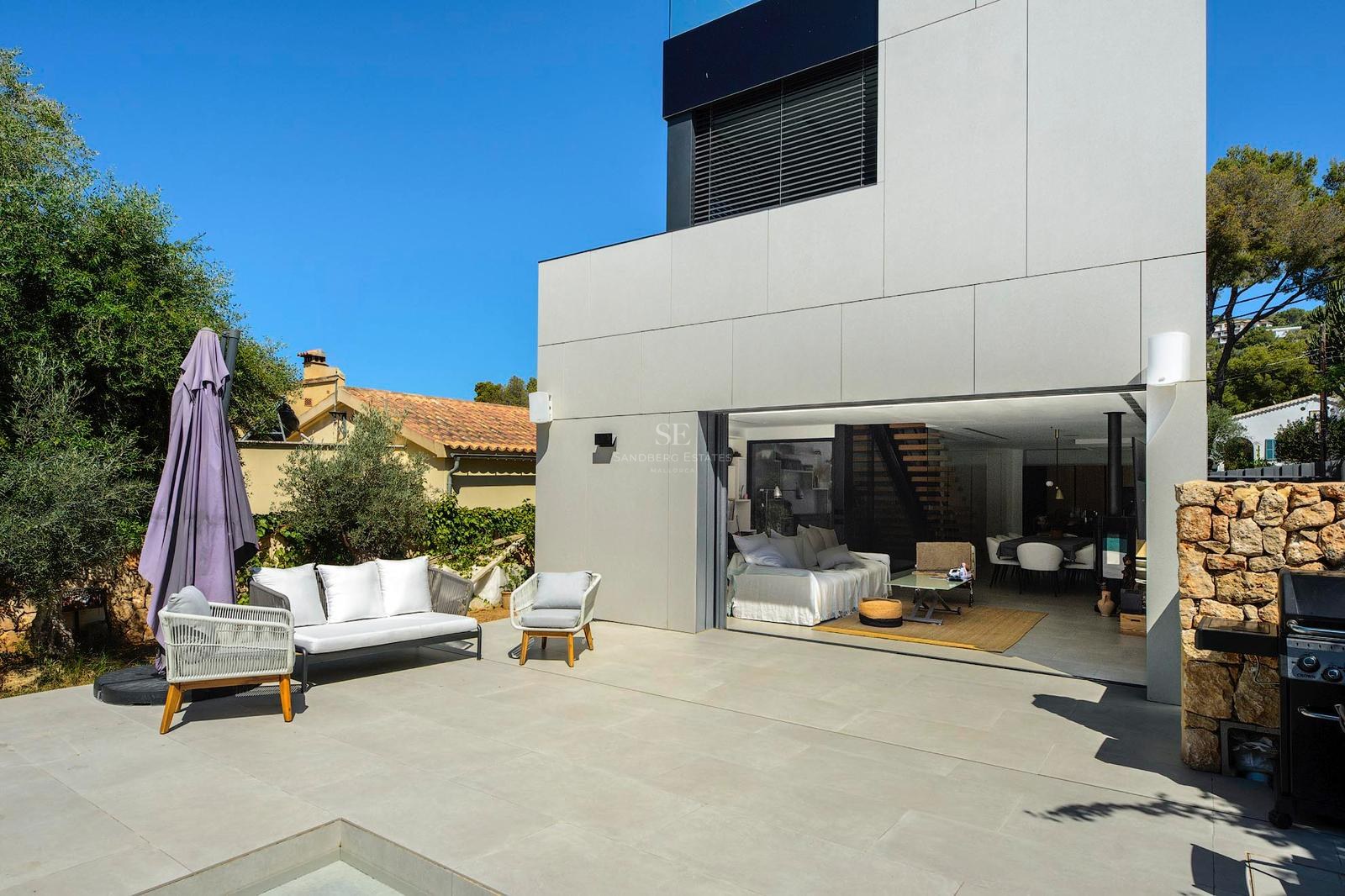 Terraza exterior moderna con muebles blancos junto a la zona de estar abierta de una villa y una pared de piedra.