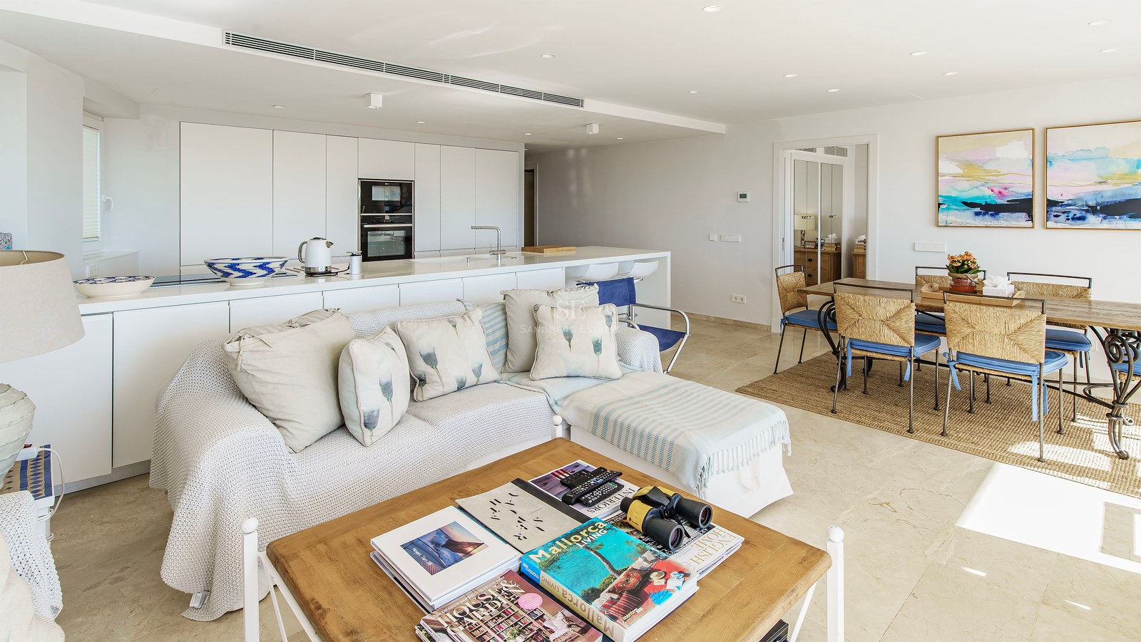 Ljust vardagsrum med öppen planlösning, vit soffa, träbord, matgrupp och minimalistiskt kök i bakgrunden.