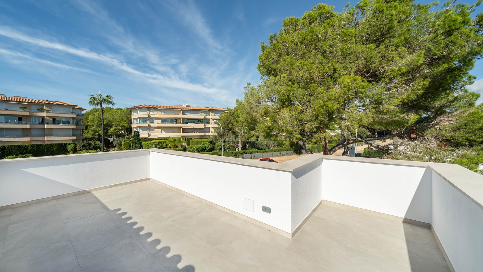 Rymlig modern terrass med vita väggar och ljusa stengolv, utsikt över frodiga tallar och byggnader.