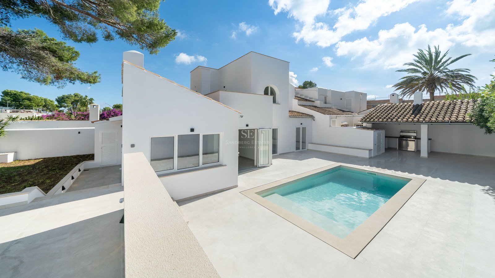 Modern white Mediterranean villa featuring a bright swimming pool, spacious stone patio, and outdoor kitchen area.