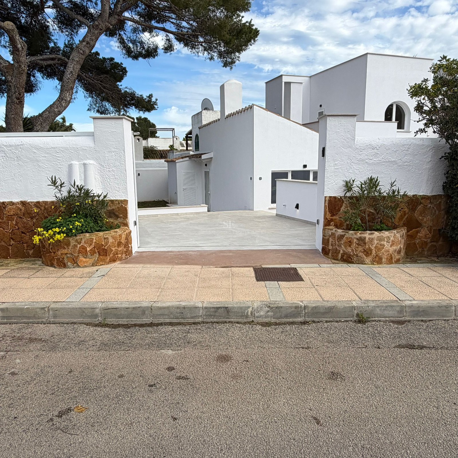 Entré till en vit villa med stenmurar, planteringar med gula blommor och stenlagd uppfart under tallar.