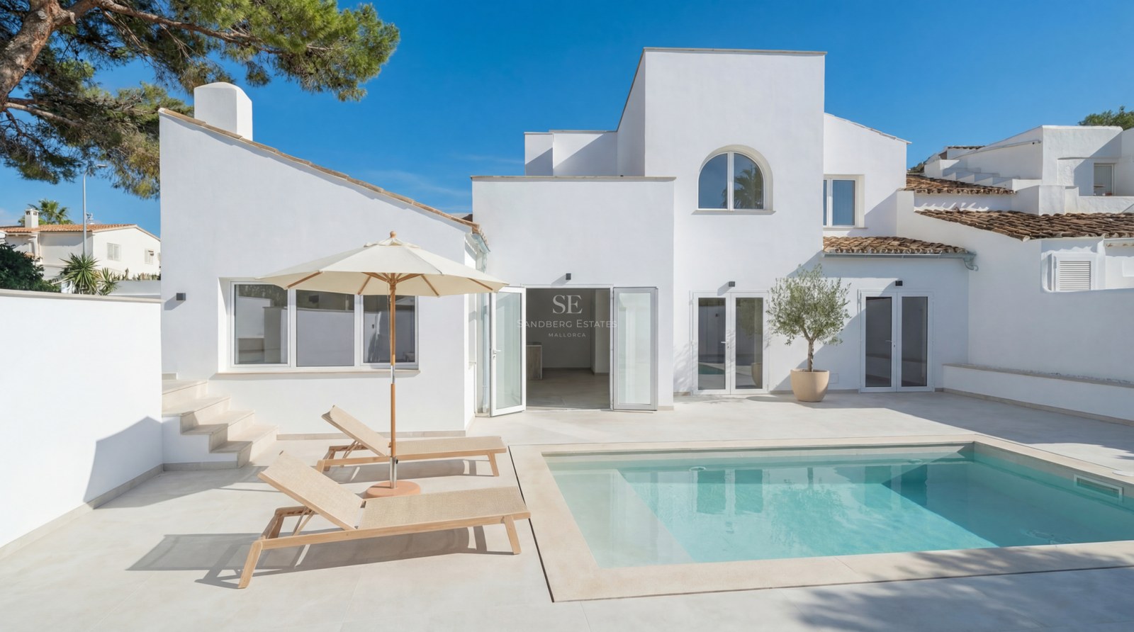 Vit modern villa med rektangulär pool, ljus stenterrass och solstolar under en klarblå himmel.