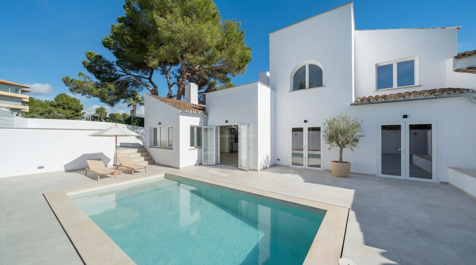 Modern vit villa med turkos pool, stenterrass och solsängar under en klarblå himmel.