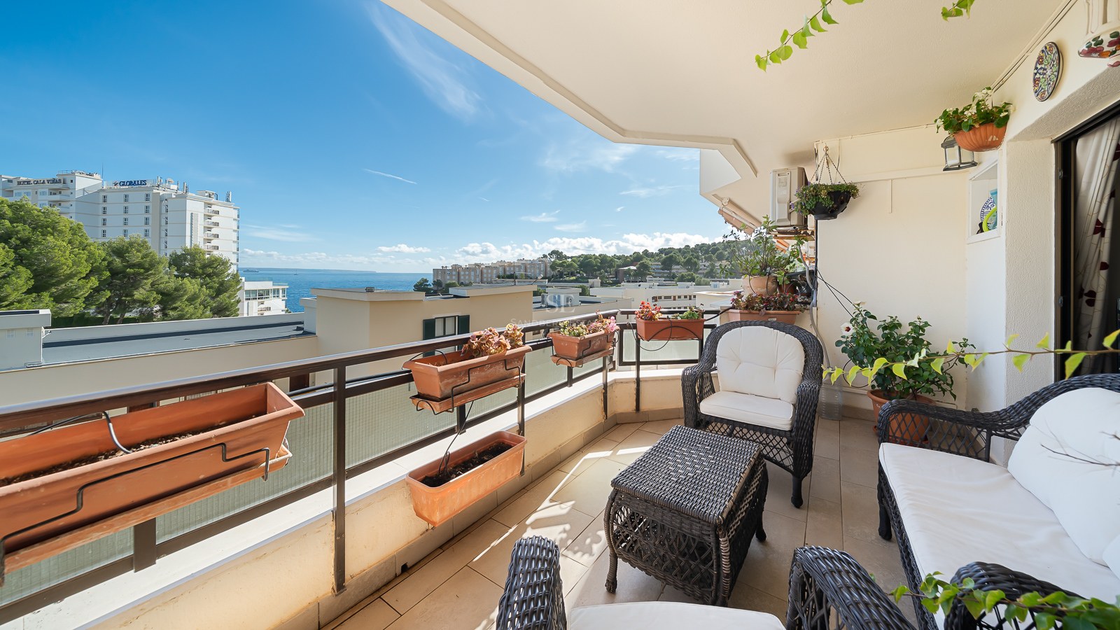Balcony with wicker furniture, white cushions, and planters overlooking a blue sea and coastal buildings.
