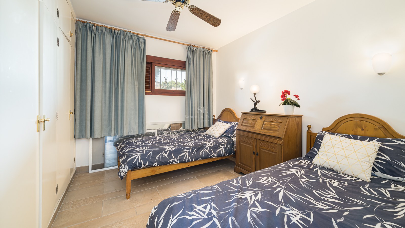 A bright bedroom featuring two wooden twin beds with blue patterned bedding, a ceiling fan, and large white wardrobes.