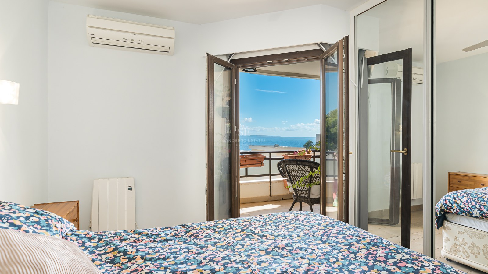 Bedroom with a floral bed overlooking a balcony with a black wicker chair and a wide view of the blue ocean.
