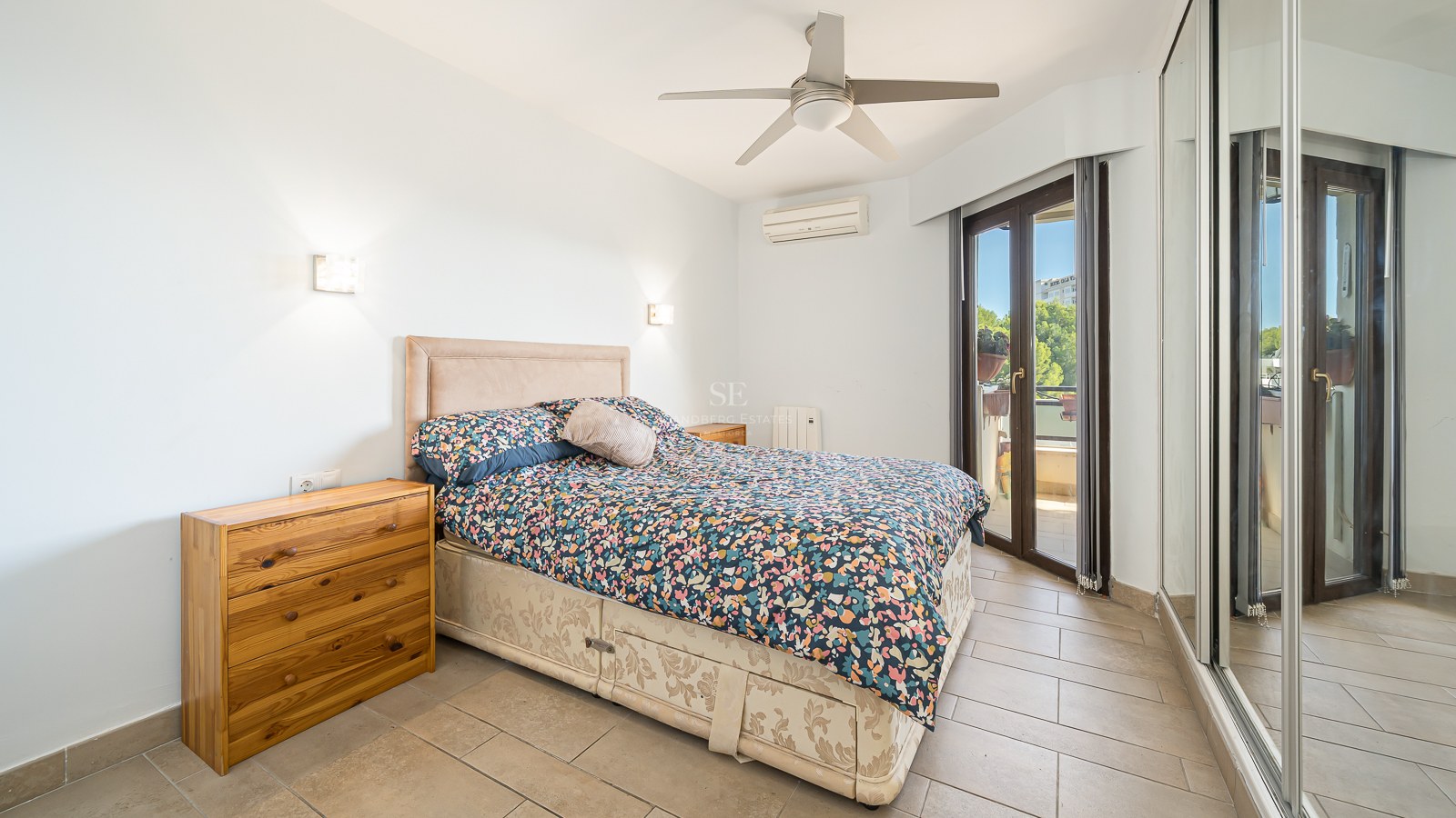 Bedroom featuring a double bed, wooden dresser, large mirrored closet, and a glass door leading to a sunlit balcony.