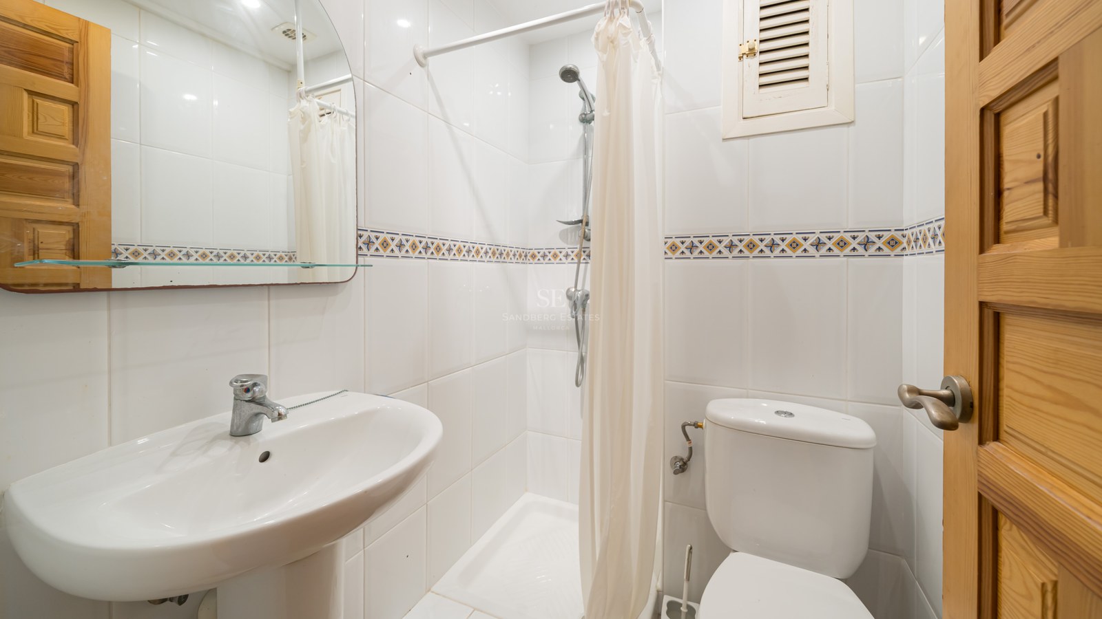 Bright bathroom with white tiles, a pedestal sink, toilet, and a walk-in shower with a white curtain.