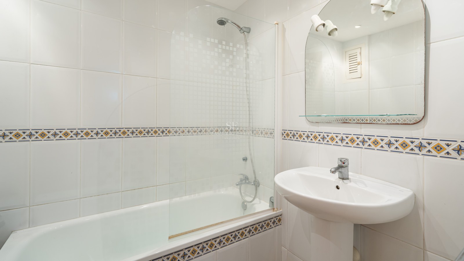 A white-tiled bathroom featuring a bathtub with a glass screen, a pedestal sink, and decorative border tiles.