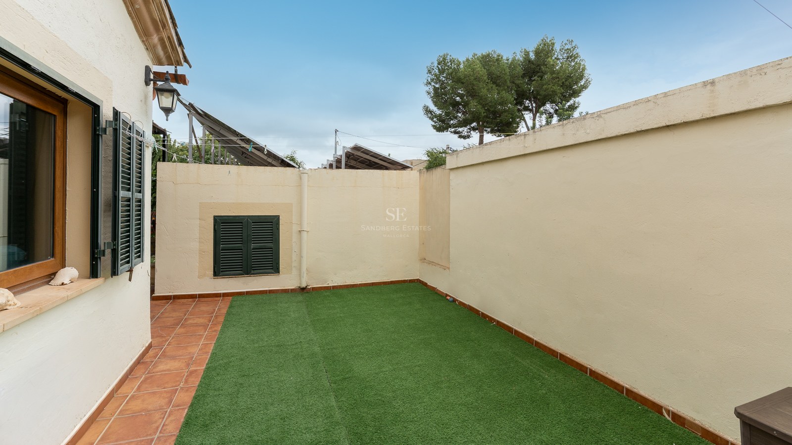 Small walled garden with artificial grass and terracotta tile border under a blue sky.