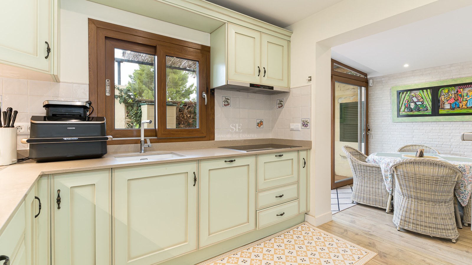 Bright kitchen with mint green cabinets, wooden window, and patterned floor leading to a dining area with wicker chairs.