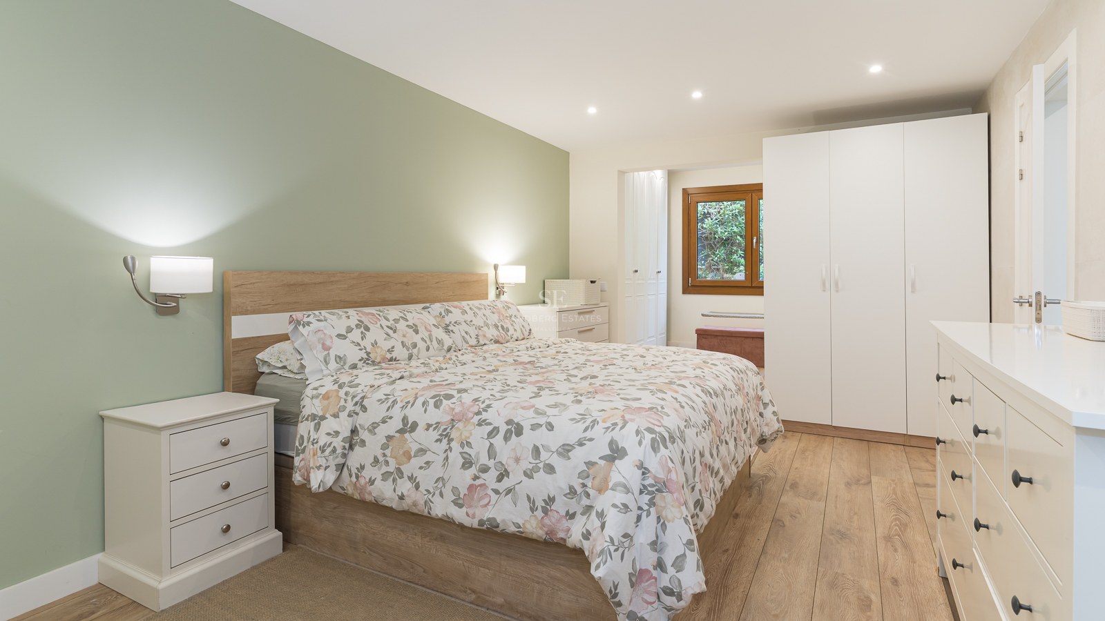 Modern bedroom featuring a floral bed, sage green wall, white furniture, and light wood flooring.